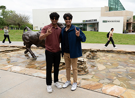 two young men give the horns up symbol in front of a bull statue outside the marshall student center